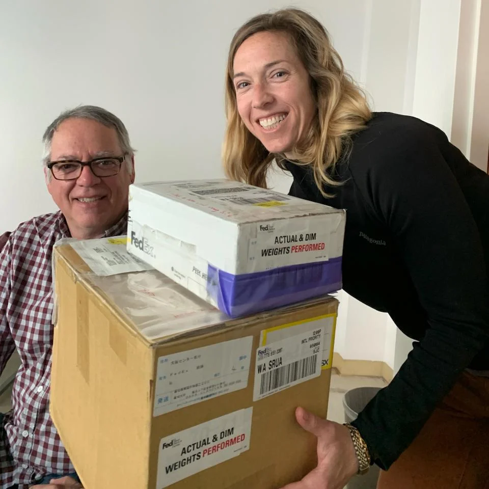 A man smiles next to a smiling woman holding a stack of cardboard boxes.