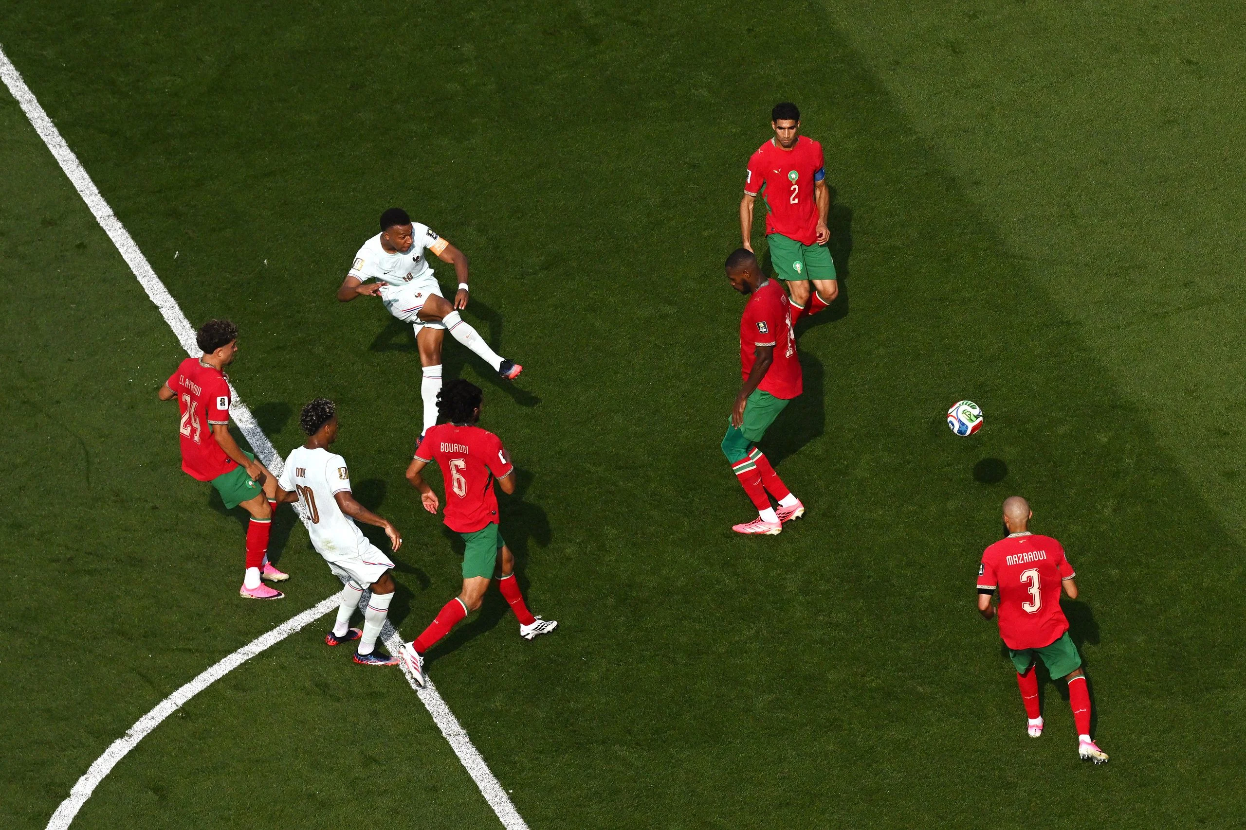 A soccer player in a white shirt kicks a ball near members of the opposing team in red shirts.