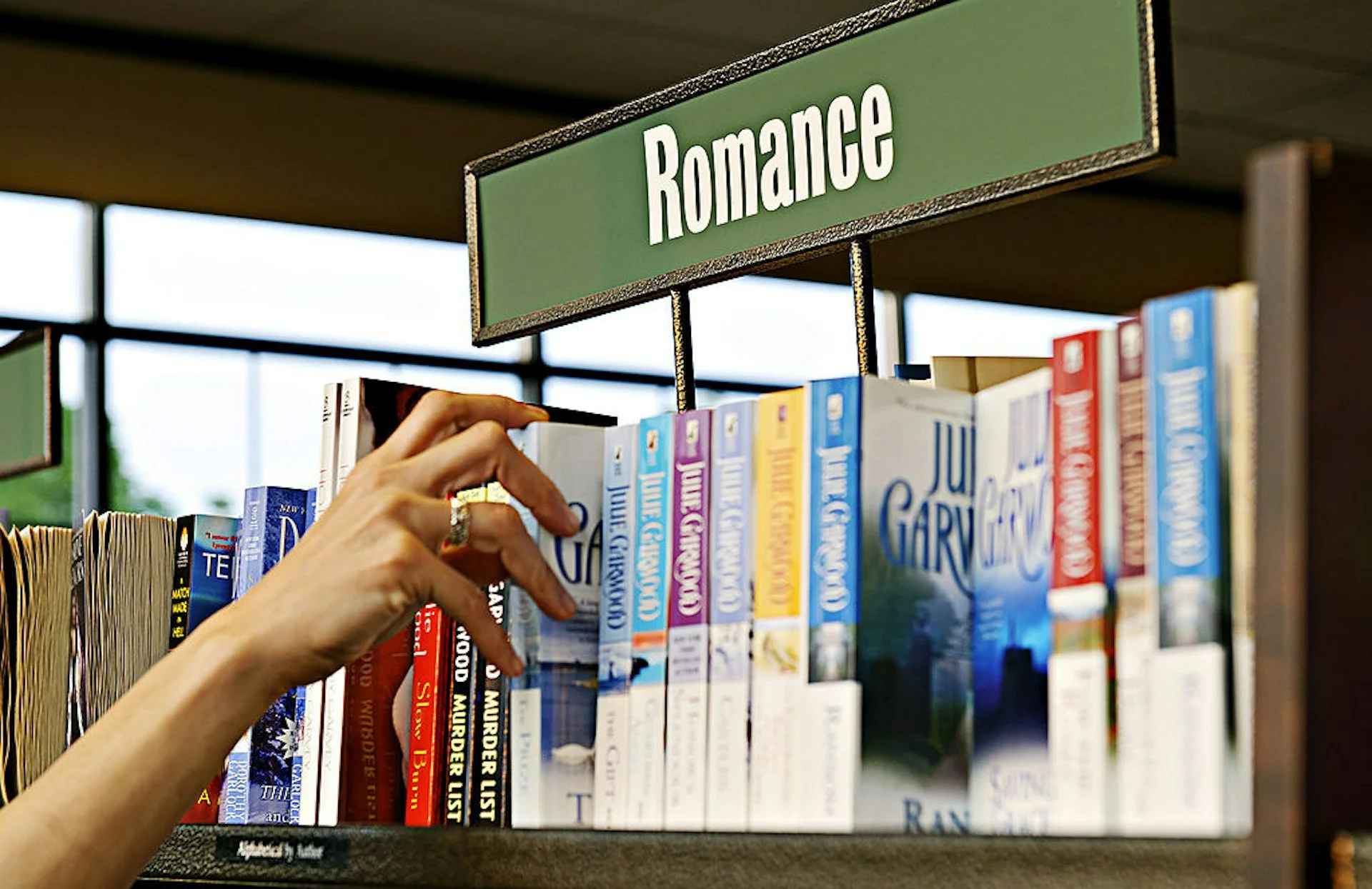 A hand reaching for a book on a shelf titled 'Romance.'