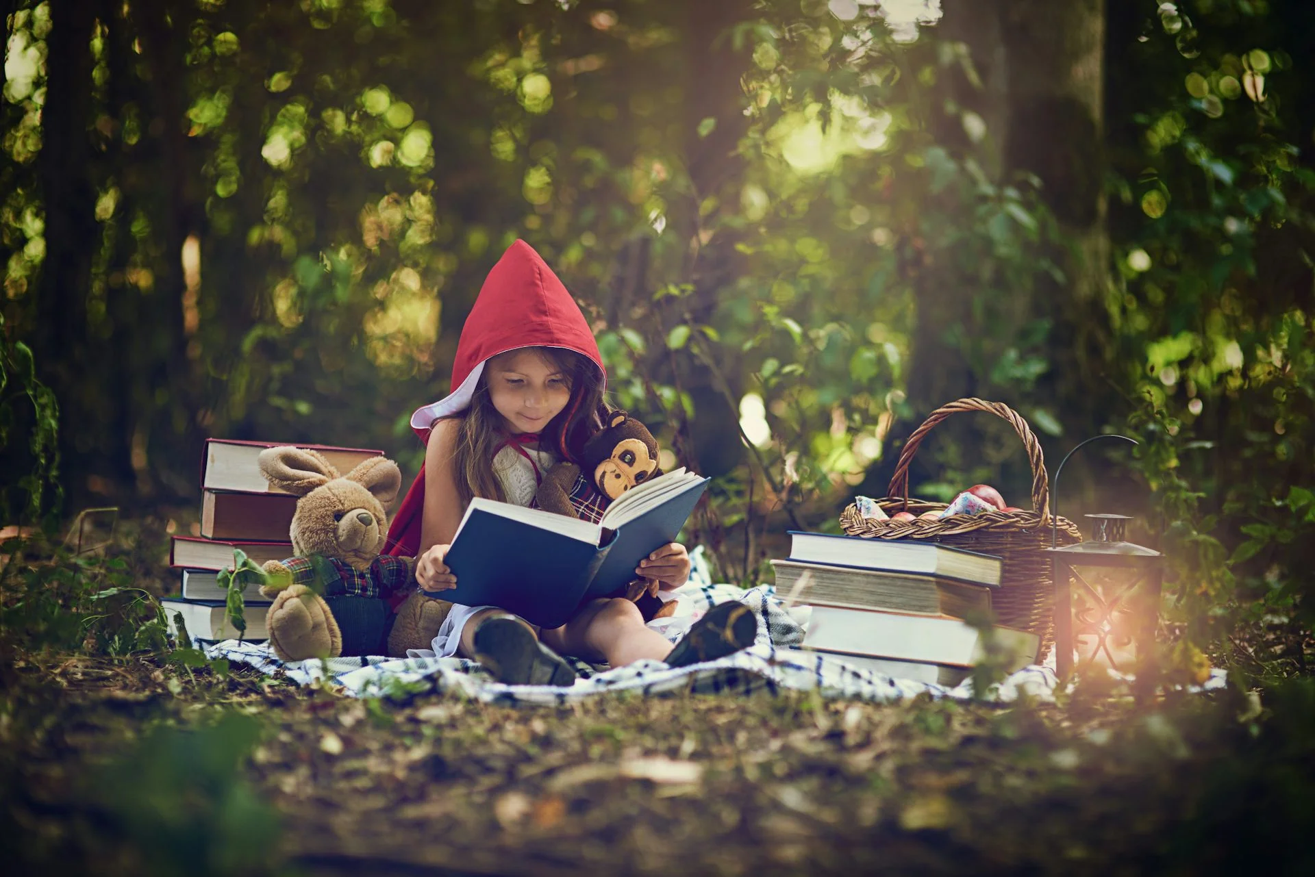 A young child wears a red pointy bonnet and looks at a book in the woods, as she sits next to two stuffed animals.