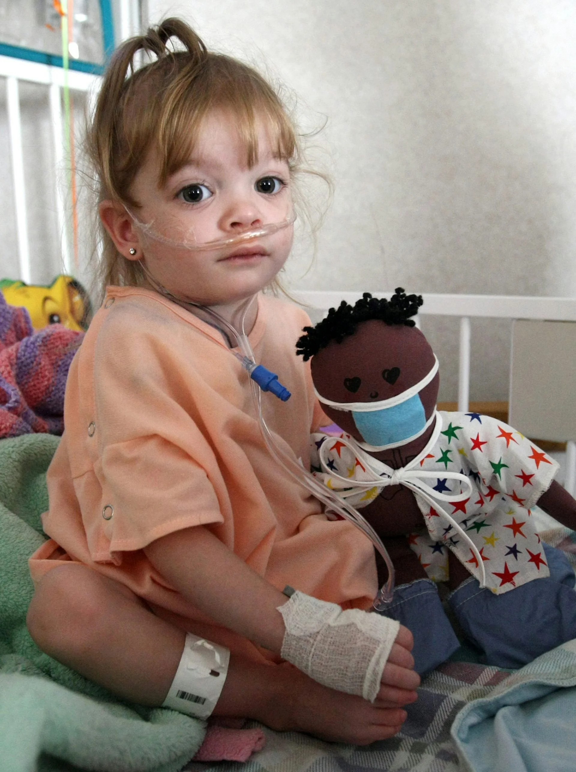 Child with nasal cannula sitting on bed, holding a doll wearing a surgical mask
