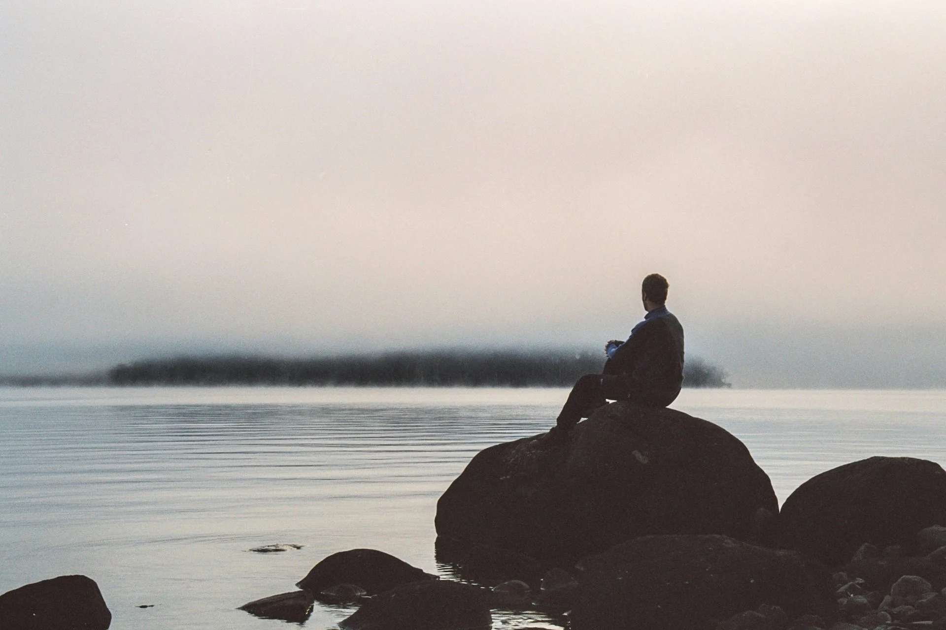 A man seen in silhouette sits atop a large rock looking out over the ocean on a gray day.